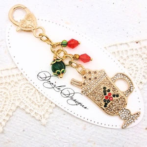 May include: Gold-toned charm with a coffee cup design, adorned with clear and red crystals, and a holly berry accent. Includes a lobster clasp and additional charms with red and green beads. The charm is attached to a white card with the text "Dazzle Designs".