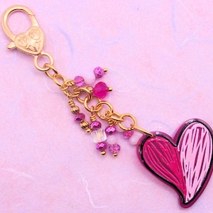 May include: A gold charm with a pink and white heart pendant and a chain of pink and clear beads. The charm is attached to a gold lobster clasp.