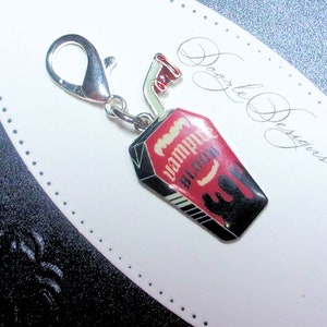 May include: A silver charm in the shape of a coffin with a red and black design. The coffin has the words "Vampire Blood" printed on it. The charm has a silver loop for attaching to a chain or bracelet.