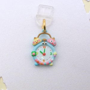 May include: A whimsical blue alarm clock charm with a gold-colored hoop. The clock face shows the time as 12:00. The charm has colorful accents and cartoon-like characters on top and sides.