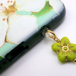 May include: A close-up of a phone case with a floral design and a green flower charm. The phone case has a white flower design on a light green background. The charm is a green flower with gold accents.