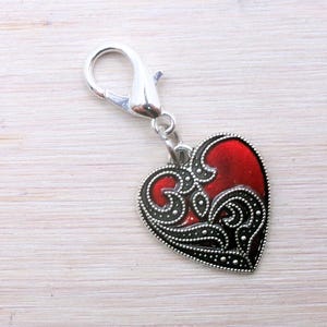 May include: A heart-shaped charm with a red enamel center and ornate black filigree design. The charm is attached to a silver-colored lobster clasp, perfect for adding to a bracelet or necklace.