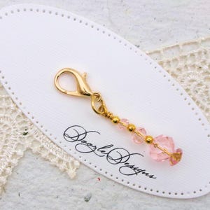 May include: A gold-colored lobster clasp charm with pink beads and gold accents. The charm is attached to a white oval tag with the text "Doozle Designs" in black script. The background includes a white lace doily.