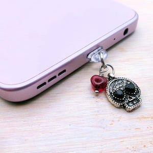 May include: A silver skull phone charm with black gem eyes and a red heart charm is attached to a light purple smartphone. The skull is detailed with small clear gems. The phone is resting on a light wood surface.