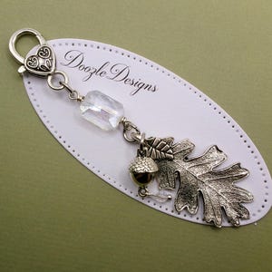 May include: A silver-toned metal keychain featuring a heart-shaped lobster clasp, a clear rectangular faceted bead, a small acorn charm, and a detailed oak leaf charm. The item is displayed on a white oval card printed with Doozle Designs.