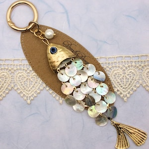 May include: A gold-colored fish-shaped keychain with iridescent, round scales. The fish has a blue eye and a pearl accent. The keychain is attached to a brown, oval-shaped tag with a gold ring.