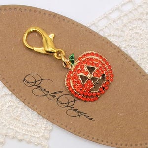May include: A gold-colored charm with a lobster clasp, featuring a pumpkin design. The pumpkin is encrusted with orange rhinestones and has triangular cutouts for eyes and a mouth. A green leaf detail is at the top.