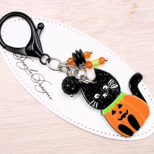May include: Black cat keychain with a pumpkin costume. The cat is wearing a green pumpkin costume with a jack-o-lantern face. The keychain has a silver chain and a black metal clasp. The keychain is on a white card with the text "Doodle Designs".