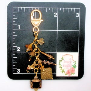 May include: A gold-colored charm with various decorative elements, including beads, a square black and gold piece, and a heart-shaped clasp. The charm is approximately 3 inches long.