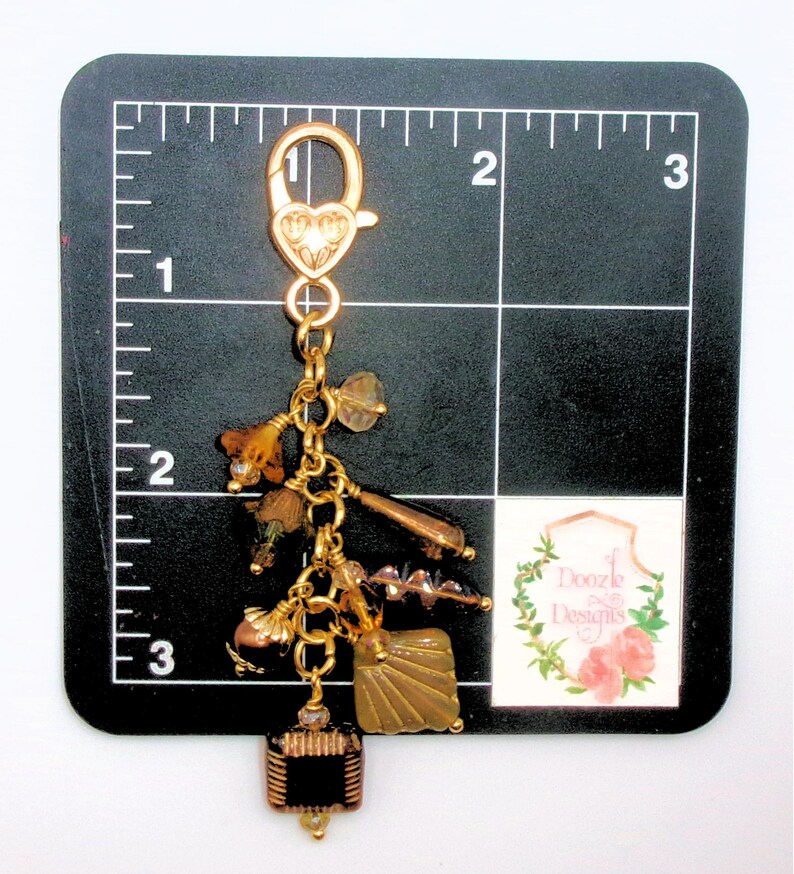 May include: A gold-colored charm with various decorative elements, including beads, a square black and gold piece, and a heart-shaped clasp. The charm is approximately 3 inches long.