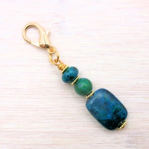 May include: A gold-colored lobster clasp charm with a string of blue and green beads. The charm features a large, rectangular blue bead at the bottom, with smaller round beads in shades of blue and green above. The beads are separated by gold-colored accents.
