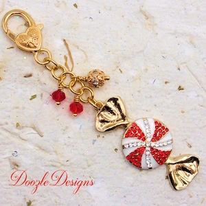 May include: A gold-tone metal keychain featuring a heart-shaped lobster clasp, a small gold bead, two red crystal charms, and a candy-shaped pendant with white enamel, red crystals, and clear rhinestones. The text Doozle Designs is visible.