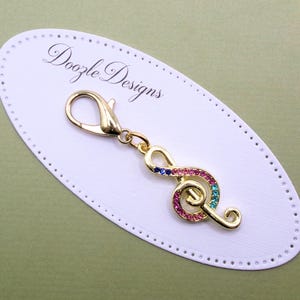 May include: A gold-toned metal treble clef charm featuring small embedded multi-colored crystals in pink, blue, and purple. The charm is attached to a gold-toned lobster clasp and is displayed on an oval white card labeled Doozle Designs.