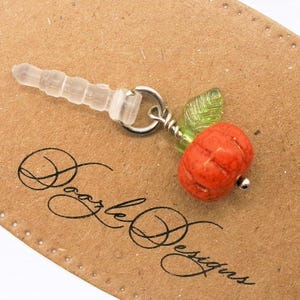 May include: A small orange pumpkin charm with a green leaf attached to a clear phone charm. The charm is attached to a silver ring.