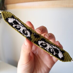 May include: Handmade friendship bracelet with a black and white pattern on a green background. The bracelet is held by a hand and has white braided cords on each end with a tassel.