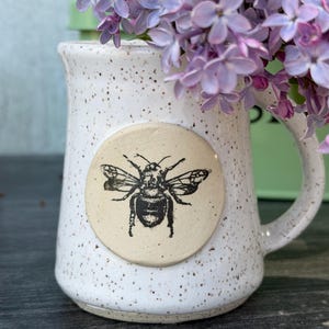 May include: A white ceramic mug with a speckled finish and a circular beige medallion featuring a black ink illustration of a bee. The mug is filled with a cluster of light purple lilac flowers, showcasing a handle on the right side.