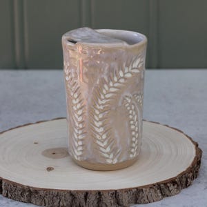 May include: A cylindrical ceramic tumbler with a glossy, light pinkish-beige glaze. The exterior features a repeating vertical pattern of white, leaf-like sprigs carved into the surface. The rim is smooth, and the base is a natural, unglazed clay.