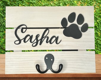 May include: A wooden pet leash holder with the name "Sasha" in black script, a black paw print, and a double hook. The background is a light wood with a green leafy backdrop.