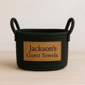 May include: Black woven basket with two handles. A rectangular brown leather patch is attached to the front, with the text "Jackson's Guest Towels" in a serif font. The basket is designed for storage.