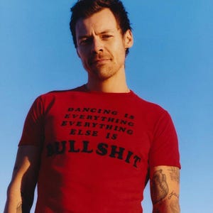 Könnte beinhalten: Ein rotes Kurzarm-T-Shirt mit schwarzem Text, der DANCING IS EVERYTHING EVERYTHING ELSE IS BULL SHIT lautet. Das Shirt hat einen Rundhalsausschnitt und wird von einer Person mit kurzem dunklem Haar und sichtbaren Tattoos an den Armen getragen.