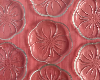 May include: A collection of clear glass plates, each shaped like a flower with petal-like edges and a central floral design. The plates are arranged on a pink surface, creating a delicate and elegant aesthetic. Ideal for serving appetizers or desserts.