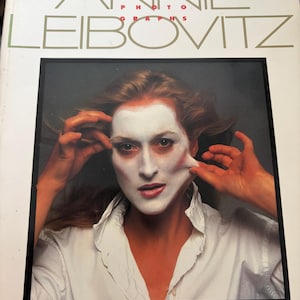 May include: A book cover featuring the title "ANNIE LEIBOVITZ PHOTOGRAPHS" in gold lettering. The cover image is a portrait of a person with white face paint and a white shirt. The introduction is by Tom Wolfe.