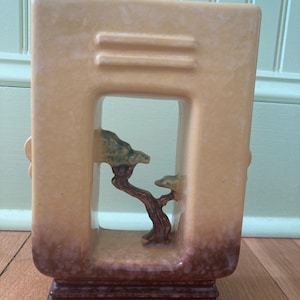 May include: A ceramic vase with a rectangular shape and a cutout design of a tree. The vase is glazed in a light brown color with a darker brown base. The tree is painted in shades of green and brown.