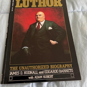 May include: A comic book titled "Lex Luthor" featuring a portrait of a man in a suit against a red background. The book's title is in gold lettering, and the cover includes the text "The Unauthorized Biography".