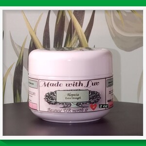 Puede incluir: Un tarro rosa con una etiqueta blanca que dice "Made with Luv Alopecia Extra Strength Because UR worth it" y un coraz&oacute;n rojo. El tarro contiene 2 oz de producto.