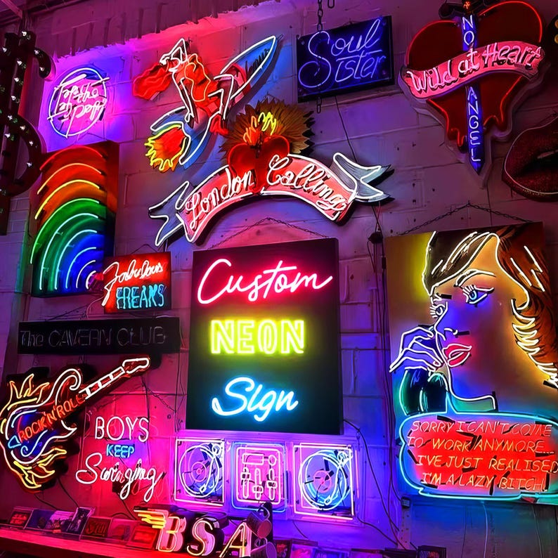 May include: A collection of vibrant neon signs mounted on a white brick wall, featuring various designs including a rainbow, a guitar, a heart, and text such as Custom Neon Sign, London Calling, Soul Sister, and Wild at Heart.
