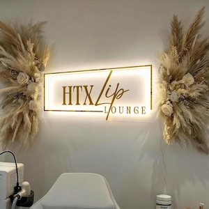 Op de afbeelding: Een goudomrand bord met de tekst "HTX Lip Lounge" is verlicht op een witte muur. Het bord wordt geflankeerd door gedroogde bloemstukken met pampasgras en witte rozen. Een witte stoel staat op de voorgrond.