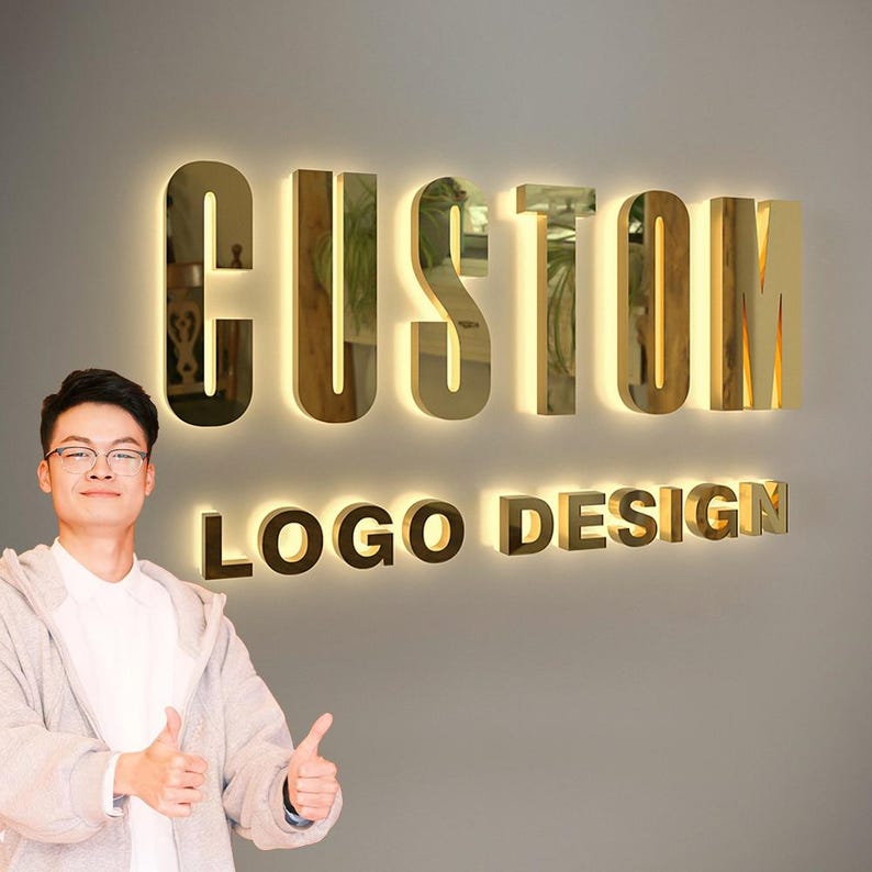 May include: Gold-colored, illuminated letters spell out "CUSTOM LOGO DESIGN" on a gray wall. The letters have a reflective surface and are lit from behind, creating a glowing effect. A person is standing in front of the sign, giving a thumbs up.