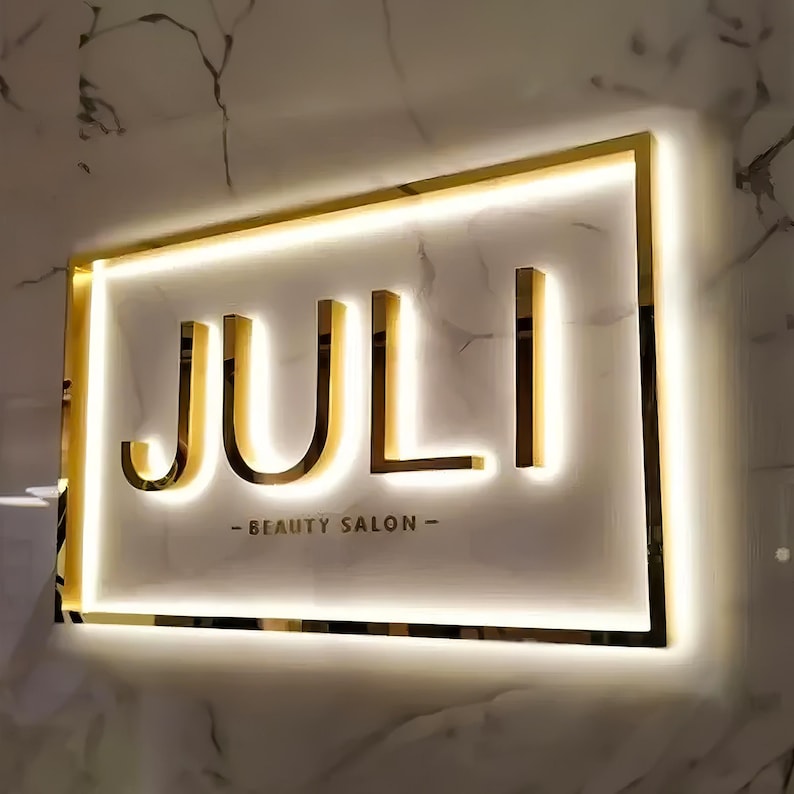 May include: A gold-colored illuminated sign for a beauty salon. The sign reads "JULI" in bold, capital letters, with "- BEAUTY SALON -" below. The sign is rectangular with a gold border and is mounted on a marble wall.