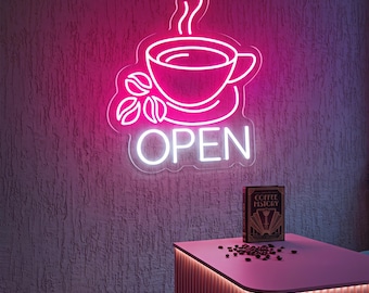 May include: A neon sign featuring a pink coffee cup with steam and three coffee beans above the white text "OPEN". The sign is mounted on a textured wall above a pink counter with a book titled "COFFEE HISTORY" and scattered coffee beans.