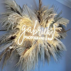 May include: A white neon sign with the text "gabriela's PHOTOGRAPHY & FILM" surrounded by dried pampas grass.