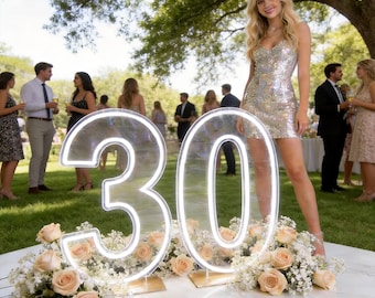 May include: A large, illuminated neon sign displaying the number 30, featuring white light tubing mounted on clear acrylic panels. The base of the sign is decorated with a dense arrangement of light peach roses and small white filler flowers.