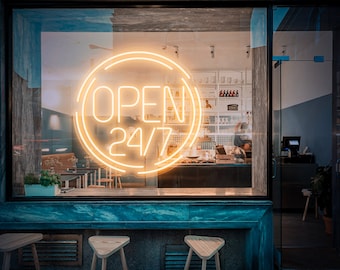 May include: A neon sign in a storefront window reads "OPEN 24/7" in a circular, yellow-orange glow. The sign is visible through a large window with a dark blue frame. Three wooden stools are in the foreground.