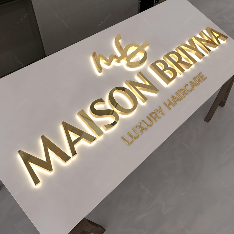 May include: A white surface with gold illuminated lettering that reads "MAISON BRYNNA LUXURY HAIRCARE". The logo features the initials "mb" in a stylized script. The letters are metallic and reflect light.