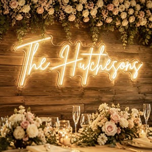 May include: A warm white neon sign featuring the script text The Hutchesons mounted on a horizontal dark brown wooden plank wall. The sign is positioned below a lush arrangement of white and pale pink roses with green foliage.