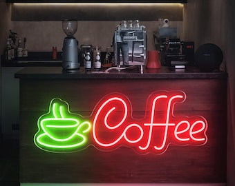May include: A red neon sign with the word "Coffee" in a cursive font. A green neon sign of a coffee cup with steam coming from it is to the left of the word "Coffee". The signs are mounted on a wooden wall.
