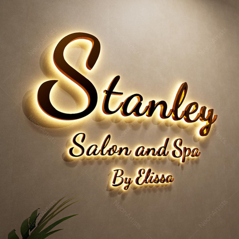 May include: A wall-mounted sign for "Stanley Salon and Spa By Elissa". The sign features a dark brown, illuminated script font against a neutral-colored wall. The letters are outlined with a warm, golden glow, creating a welcoming ambiance.