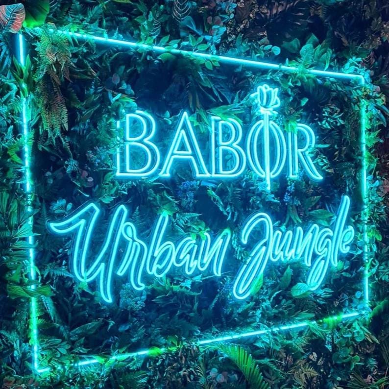 May include: A rectangular neon sign with bright blue light emitting the text BABOR and Urban Jungle, featuring a stylized rose icon above the text, all mounted against a dense background of various green artificial tropical leaves.