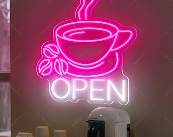 May include: A neon sign in the shape of a coffee cup with steam, coffee beans, and the word "OPEN" in white. The sign is a bright pink color and is mounted on a wall. Below the sign is a white coffee machine and cups.