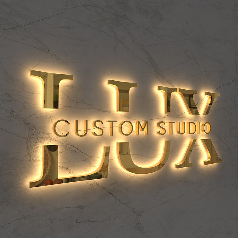 May include: Gold-colored illuminated sign on a marble wall. The sign reads "TIV CUSTOM STUDIO LUA" in a stylized font. The letters are three-dimensional and cast a warm glow, creating a luxurious aesthetic.