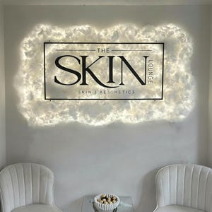 May include: A wall-mounted sign for "The Skin Lounge" with a black frame and white text. The sign is surrounded by a cloud-like, illuminated border. Two white chairs are visible below the sign.