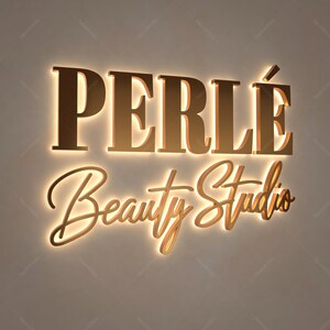 May include: A gold-colored sign with the words "PERLE Beauty Studio" in a stylish font. The sign is illuminated with a warm, inviting glow, creating a luxurious and elegant aesthetic. The sign is likely for a beauty salon or spa.
