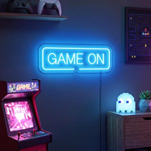 May include: A rectangular neon sign featuring the text GAME ON in bright blue light. The sign has a rounded border and is mounted on a dark wall. It emits a vibrant glow, illuminating the surrounding space with a cool blue hue, creating a distinct visual focal point in the room.