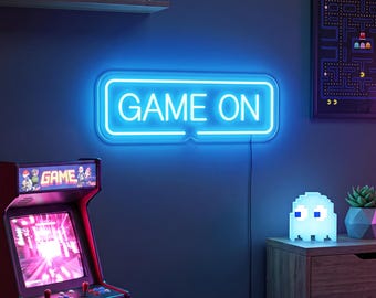 May include: A rectangular neon sign featuring the text GAME ON in bright blue light. The sign has a rounded border and is mounted on a dark wall. It emits a vibrant glow, illuminating the surrounding space with a cool blue hue, creating a distinct visual focal point in the room.