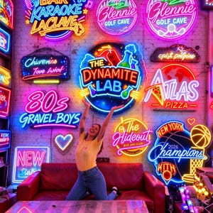 May include: A wall covered in various colorful neon signs including text such as Bar Karaoke LaClave, The Dynamite Lab, Atlas Pizza, 80s Gravel Boys, and New York Champions 2026, positioned above a red leather sofa and a rectangular table.