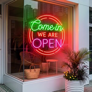 Pode incluir: Um letreiro de néon circular numa montra exibe o texto Come in WE ARE OPEN em luz verde, laranja e rosa. A exposição inclui uma cadeira de arame dourada, um saco de palha entrançado, uma pequena mesa lateral branca e um vaso branco canelado.